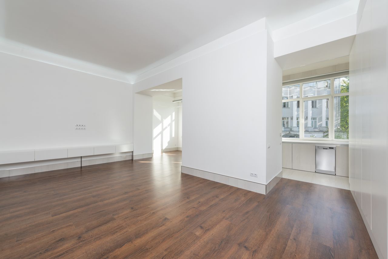 services-05 A contemporary empty apartment with white walls and wooden flooring, featuring large windows.